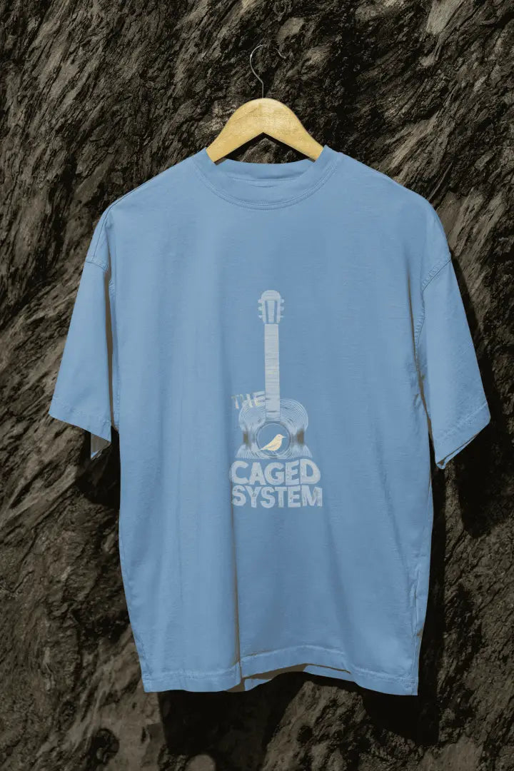 A baby blue oversized t-shirt with a minimal design printed on the front hanging against a tree trunk. The design consists of an acoustic guitar with a bird sitting inside it. There's a text saying The Caged System at the bottom of the design.