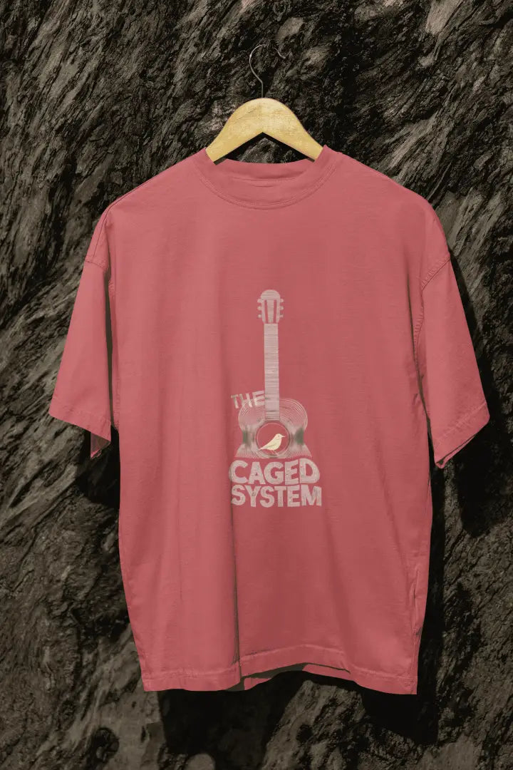 A dusty rose oversized t-shirt with a minimal design printed on the front hanging against a tree trunk. The design consists of an acoustic guitar with a bird sitting inside it. There's a text saying The Caged System at the bottom of the design.