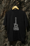 A black oversized t-shirt with a minimal design printed on the front hanging against a tree trunk. The design consists of an acoustic guitar with a bird sitting inside it. There's a text saying The Caged System at the bottom of the design.