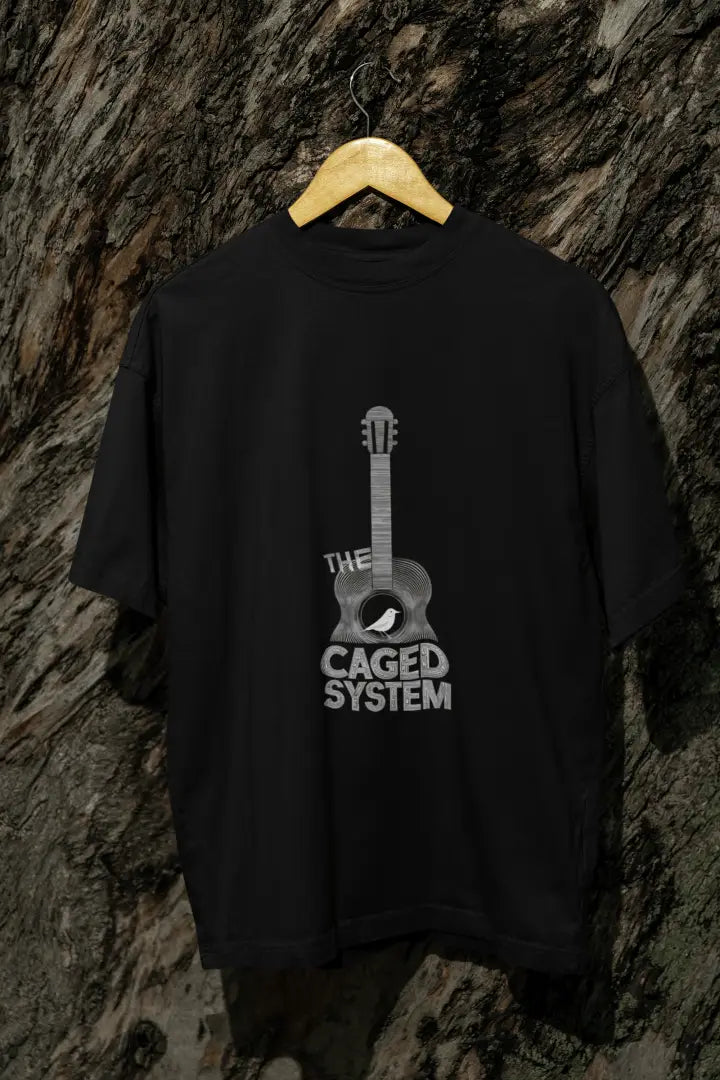 A black oversized t-shirt with a minimal design printed on the front hanging against a tree trunk. The design consists of an acoustic guitar with a bird sitting inside it. There's a text saying The Caged System at the bottom of the design.