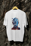 A white oversized t-shirt with a design printed at the back hanging against a tree trunk. The design contains a character whose face is made out of ripples of wave. There's a text saying Timeless Ripples at the center of the design.
