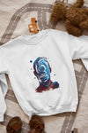 A white sweatshirt with a design printed on the front lying on the floor. The design contains a character whose face is made out of ripples of wave. There's a text saying Timeless Ripples at the center of the design.